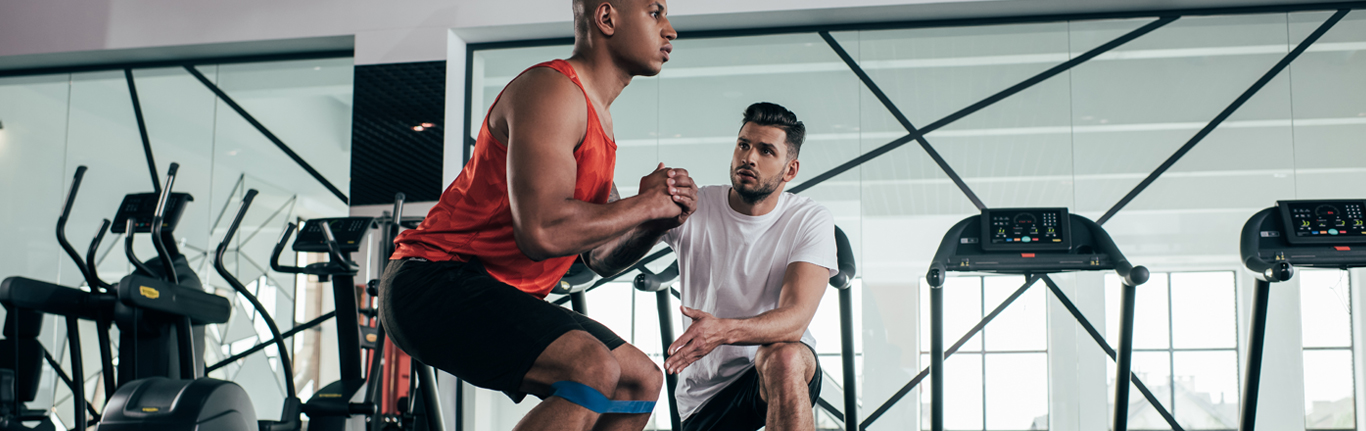 Attentive trainer looking at young African American sportsman warming up in gym.