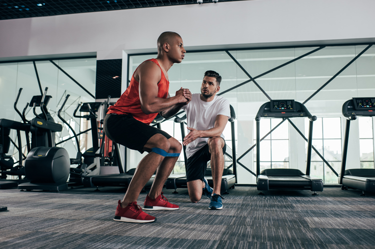 Attentive trainer looking at young African American sportsman warming up in gym.