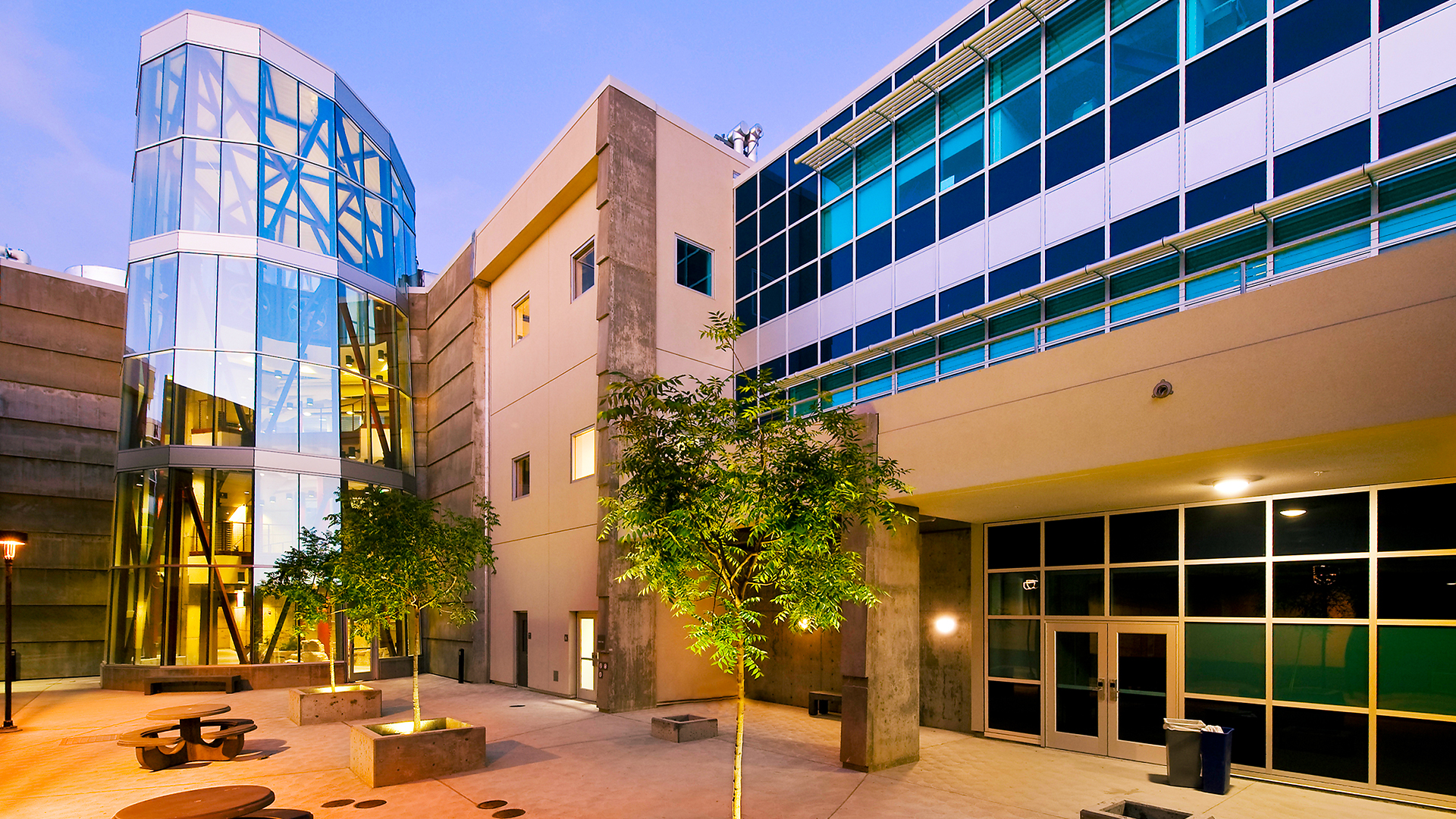Modern architecture of a building courtyard featuring glass and concrete structures with trees and seating areas.