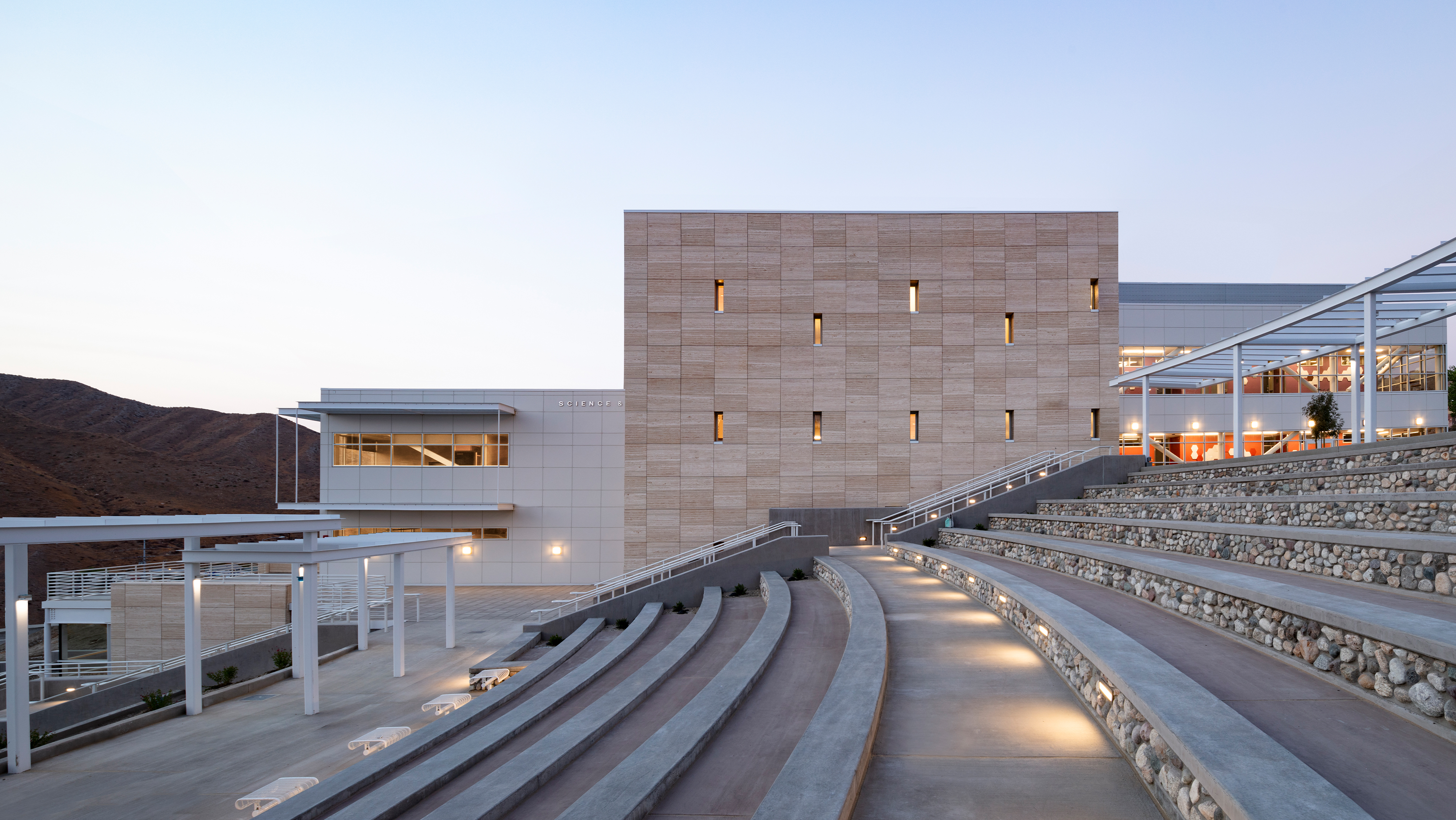 Modern outdoor amphitheater with tiered seating, leading to a contemporary building set against a mountainous landscape at dusk.