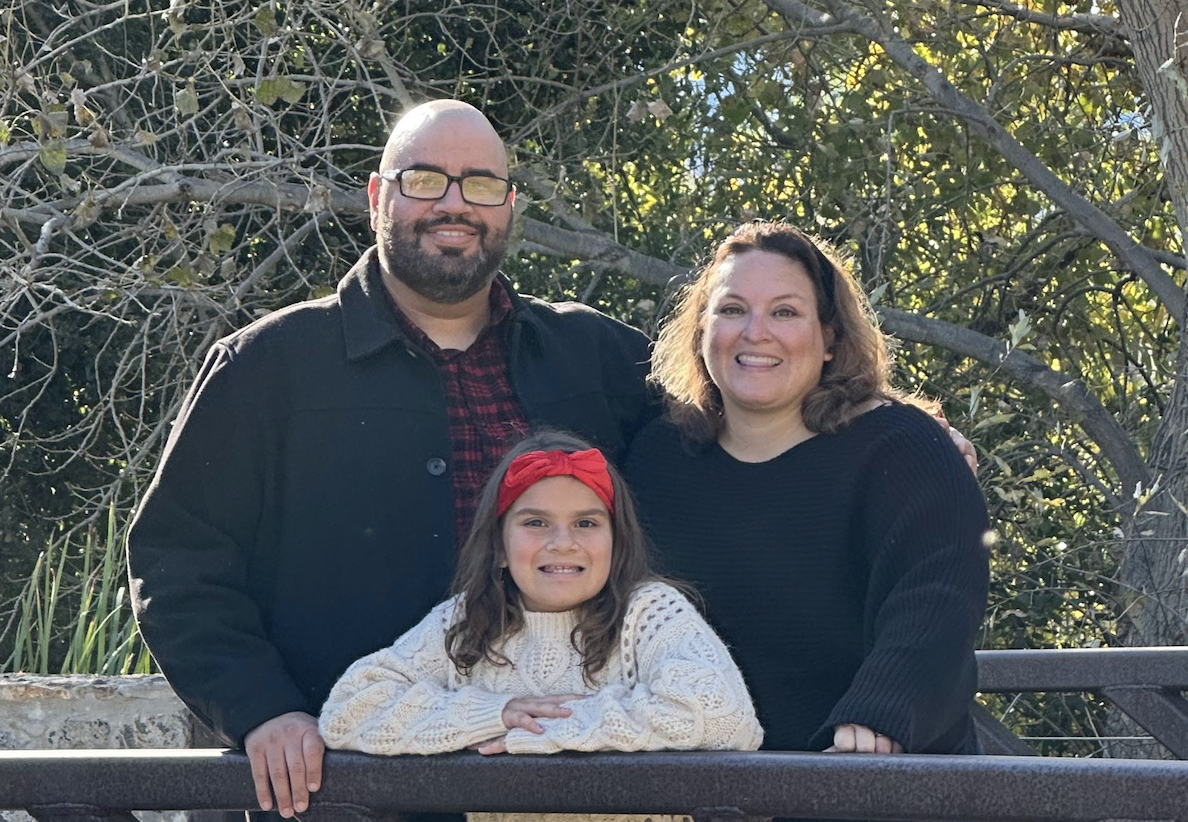 A family photo on a bridge, two parents and one child surrounded by trees.