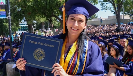 A female student in cap and gown smiles while holding up her degree during the commencement ceremony in the Honor Grove. Fellow graduates are seated behind her.