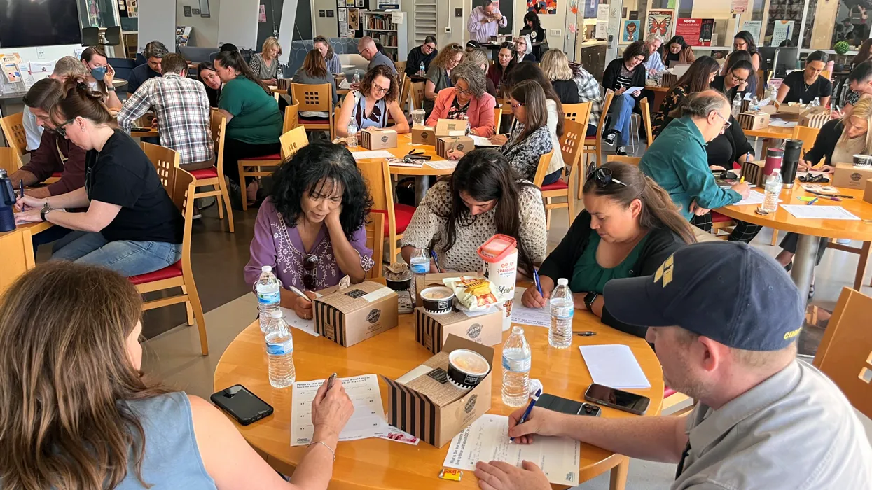 Group of adults seated at round tables in a bright community room, writing on worksheets with lunch boxes and water bottles on the tables.