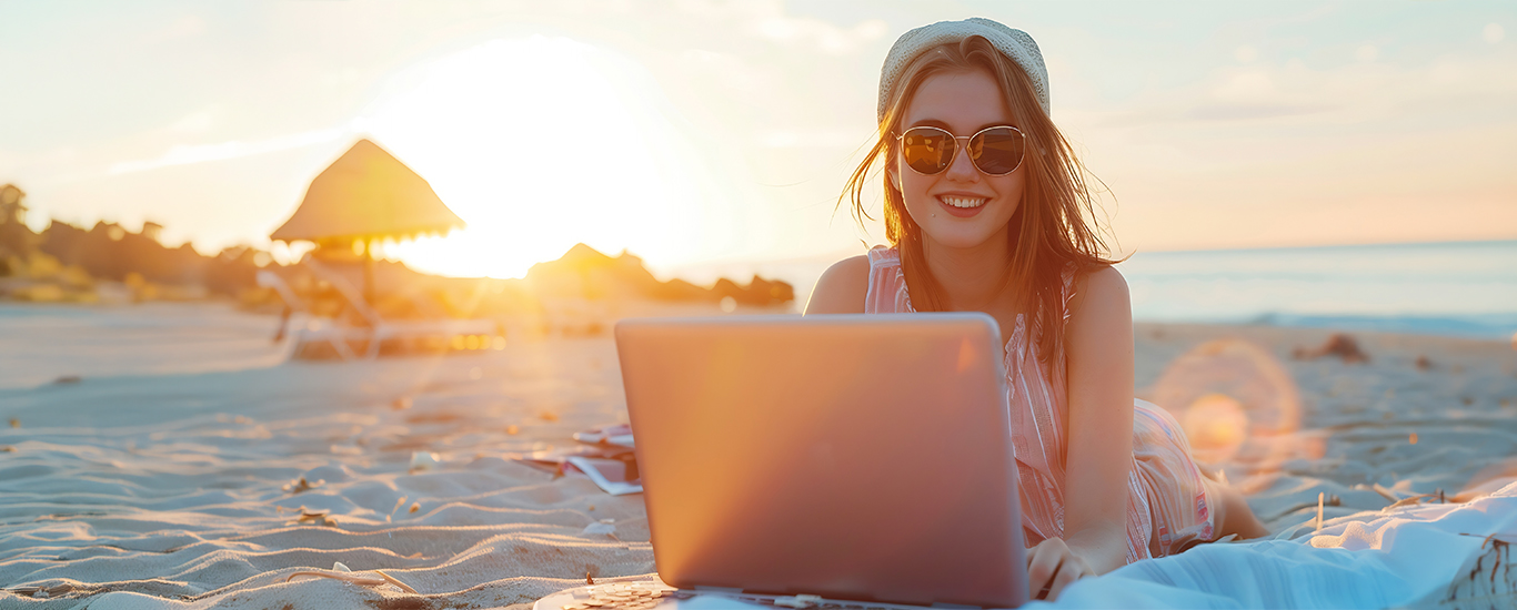 A young woman holds a laptop computer while laying on a sandy beach. 