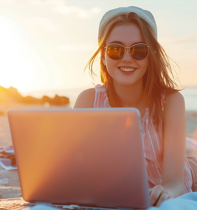 A young woman holds a laptop computer while laying on a sandy beach. 