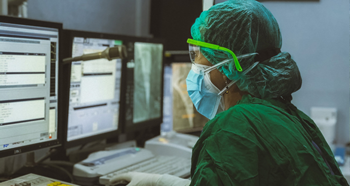 A nurse in scrubs and protective gear looks at data on a computer screen.