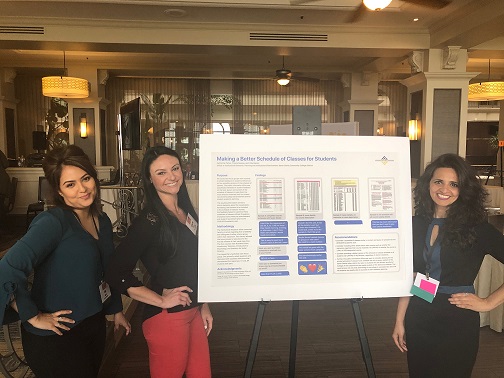 Three presenters, from left to right, Vida Manzo, Catherine Parker, and Preeta Saxena, stand posing with their hands on their hips with a poster made by a Graphic Multimedia Design student titled "Making a Better Schedule of Classes for Students"