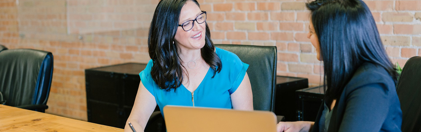 Two women seated and talking at a table with a laptop.