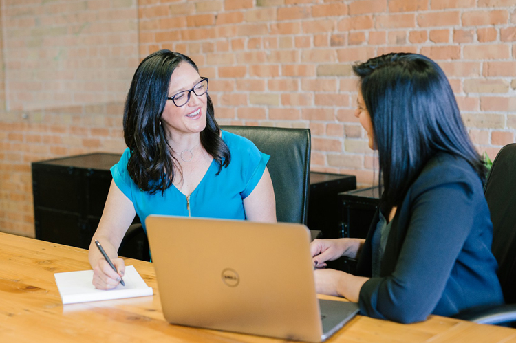 Two women seated and talking at a table with a laptop.